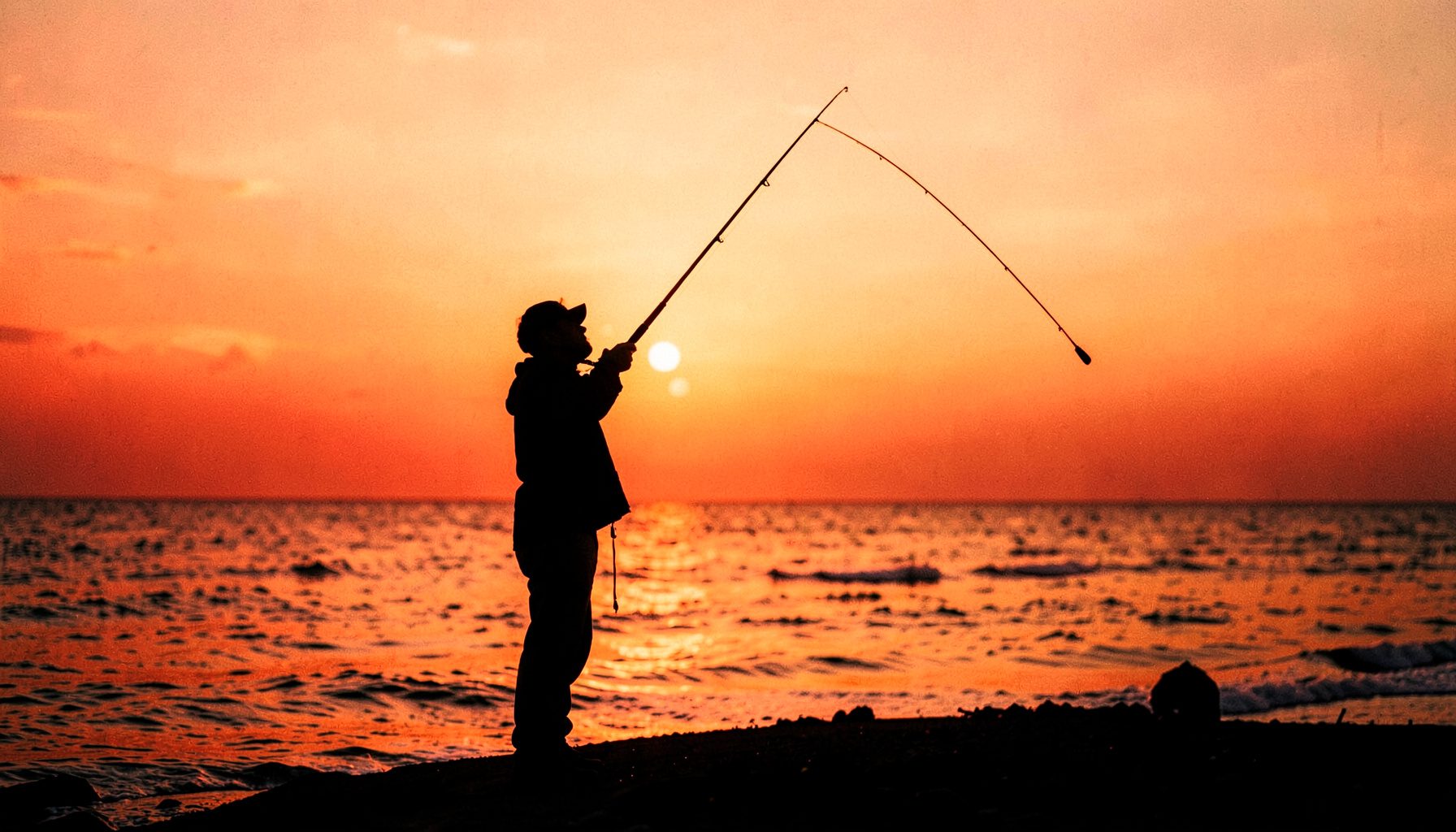 Pescador de pie en la orilla al amanecer, con la caña en posición de lanzamiento, silueta recortada contra un cielo anaranjado y dorado.