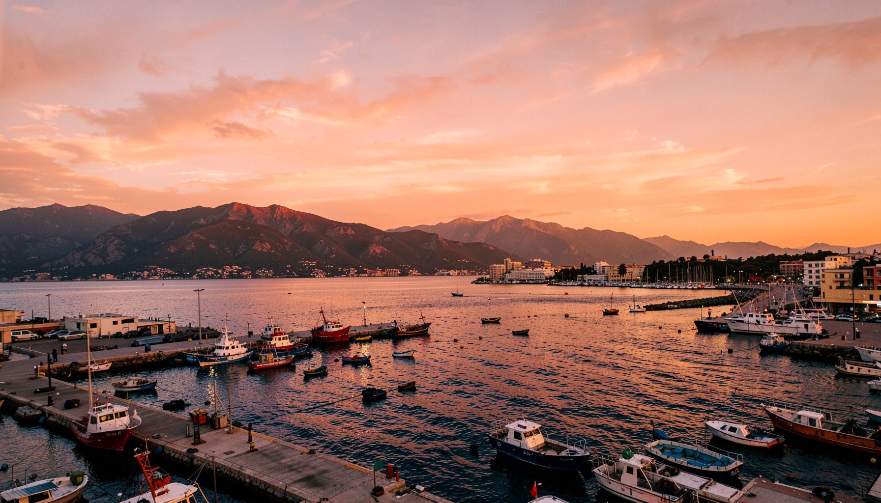 Vista panorámica del puerto deportivo de Adra al atardecer, con el mar Mediterráneo en tonos azul profundo y dorado, montañas al fondo y embarcaciones de pesca amarradas en los pantalanes. Cielo amplio con nubes suaves anaranjadas.
