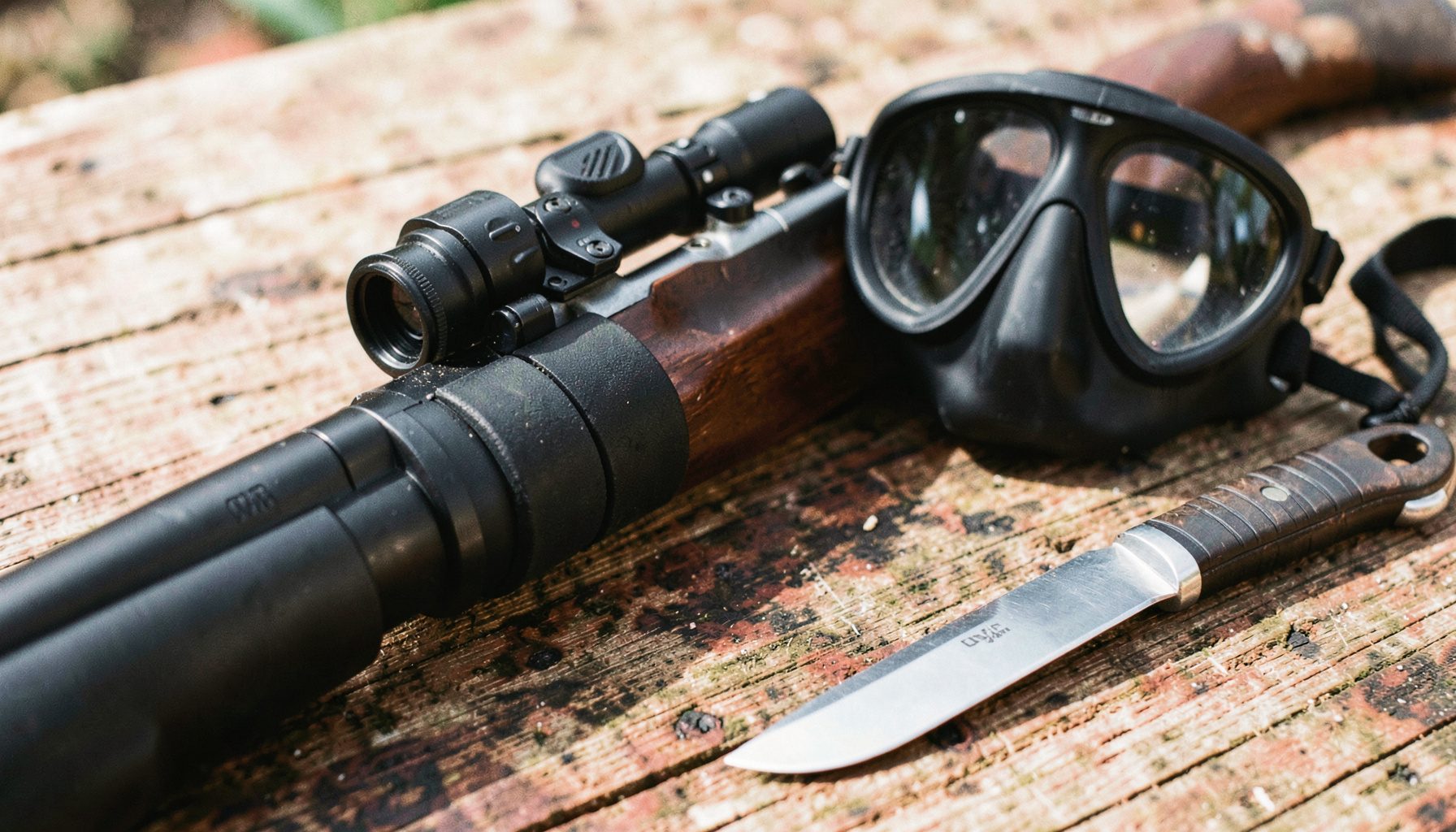 Primer plano de un fusil de pesca submarina con bandas de goma negras sobre una superficie de madera desgastada, junto a una máscara de buceo de silicona negra y un cuchillo de acero inoxidable con funda, iluminados por luz natural suave.