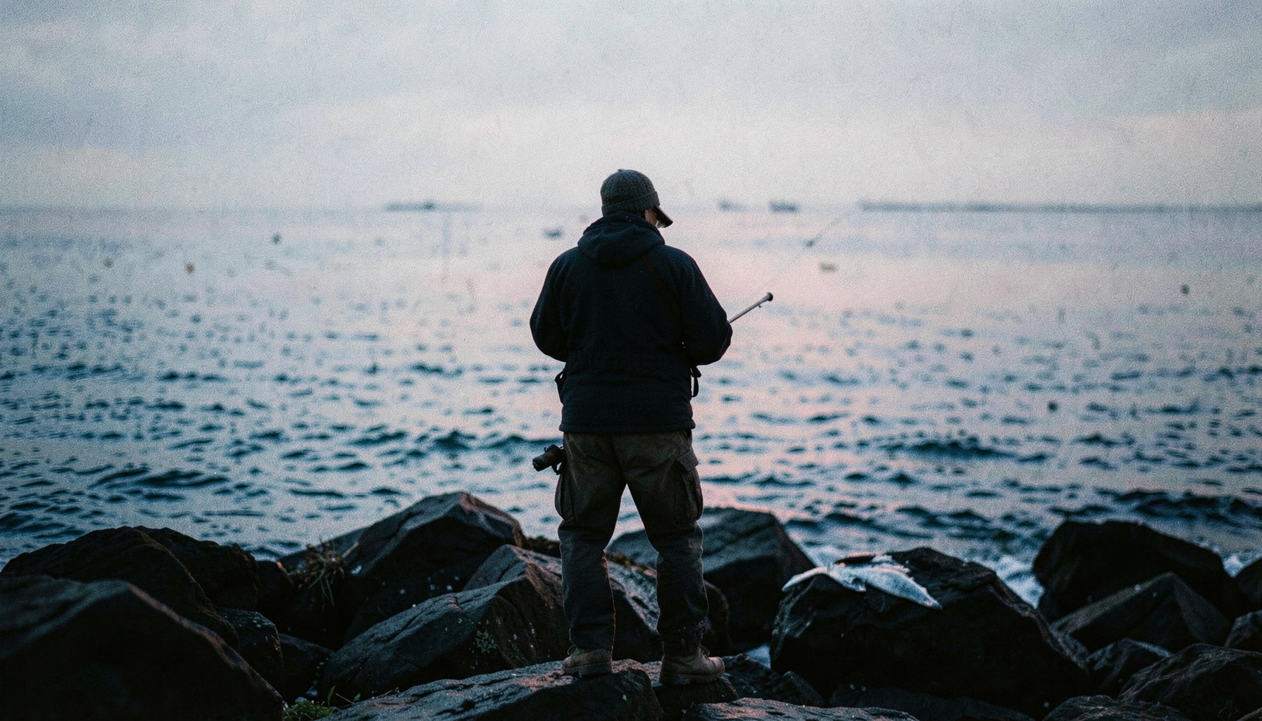 Fotografía documental de un pescador visto de espaldas al amanecer sobre rocas junto al mar, con tonos azules y grises apagados y un ambiente tranquilo y contemplativo